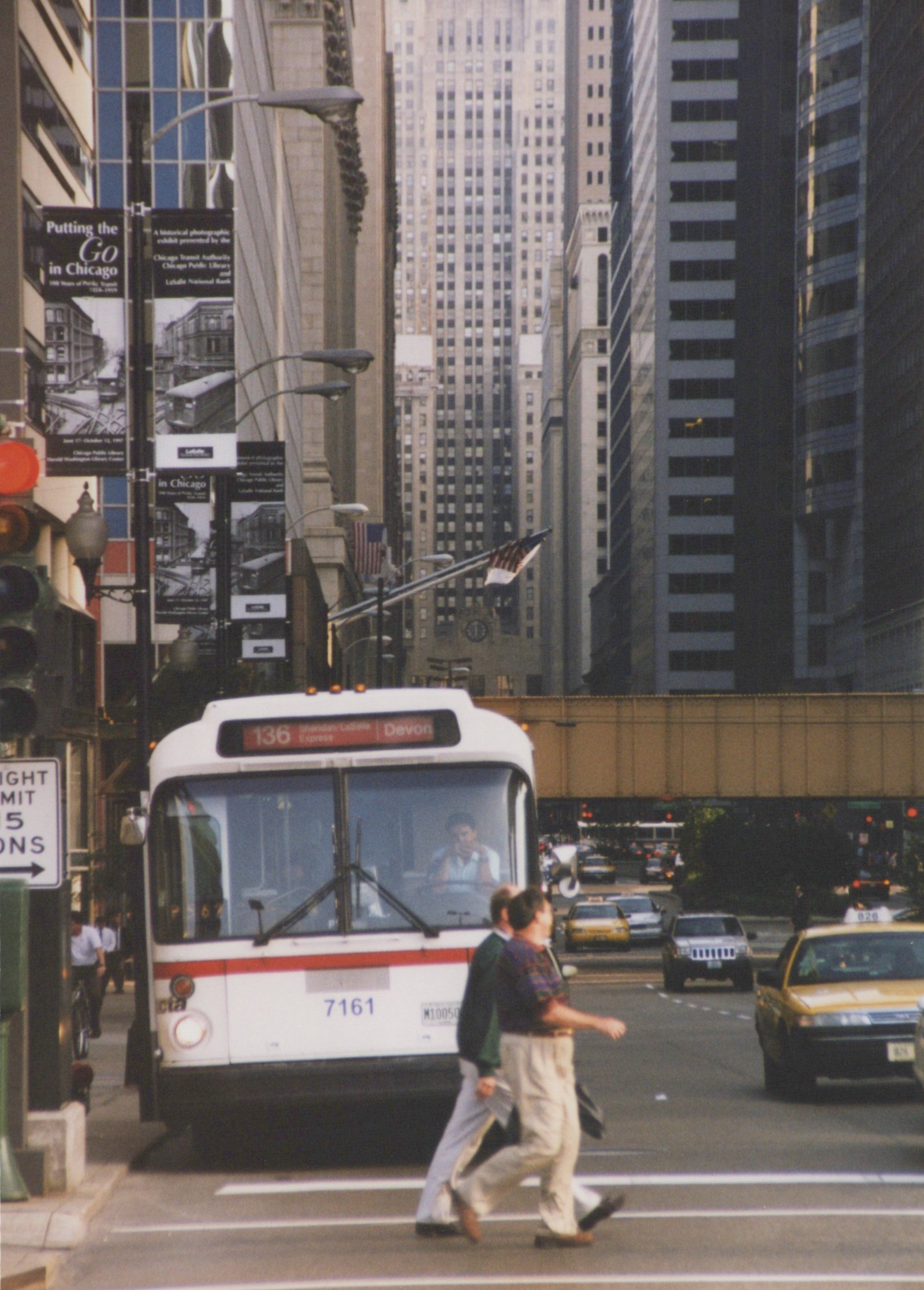 Front view of a white and red CTA bus numbered 7161 on a city street, with people crossing in front and skyscrapers in the background.