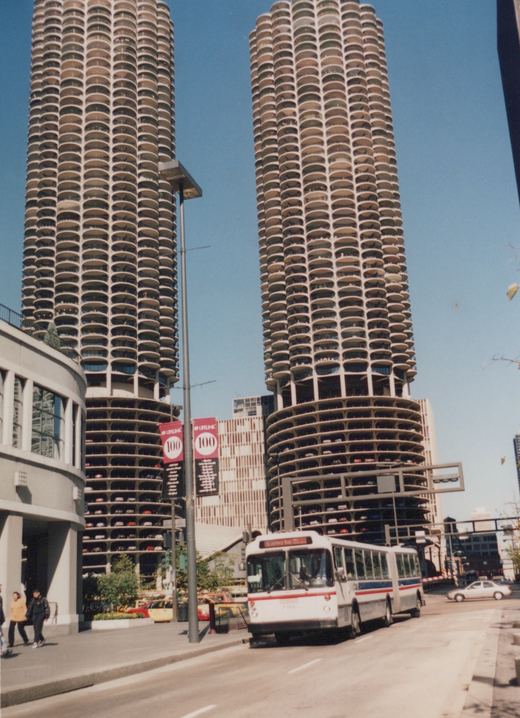 A white, red, and blue Chicago Transit Authority bus, number 7166, is stopped on a city street in front of the distinctive cylindrical Marina City towers on a sunny day.