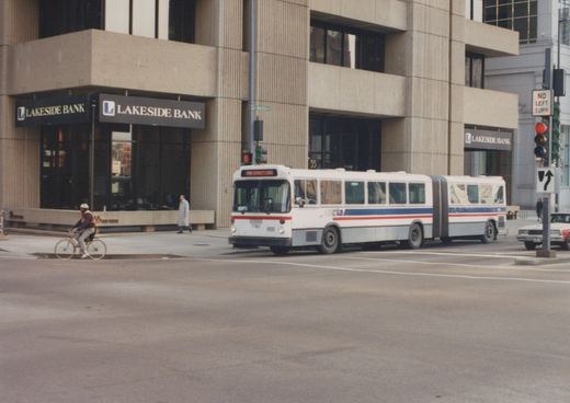 A white and blue Chicago Transit Authority articulated bus with the number 7161 on the front is stopped at a street intersection with a cyclist and a pedestrian nearby. The background shows a building with a Lakeside Bank sign and a traffic signal.