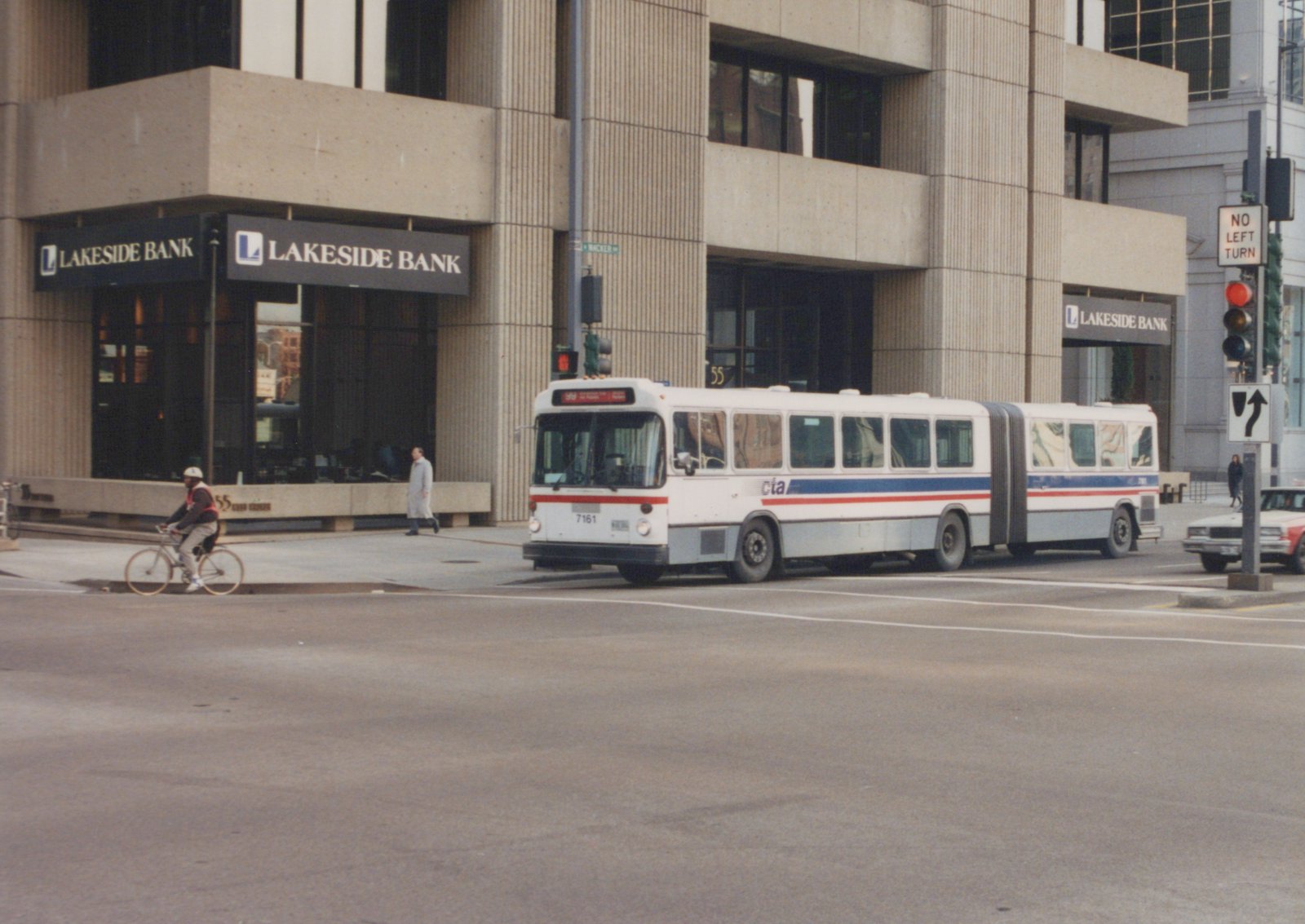 A white and blue Chicago Transit Authority articulated bus with the number 7161 on the front is stopped at a street intersection with a cyclist and a pedestrian nearby. The background shows a building with a Lakeside Bank sign and a traffic signal.