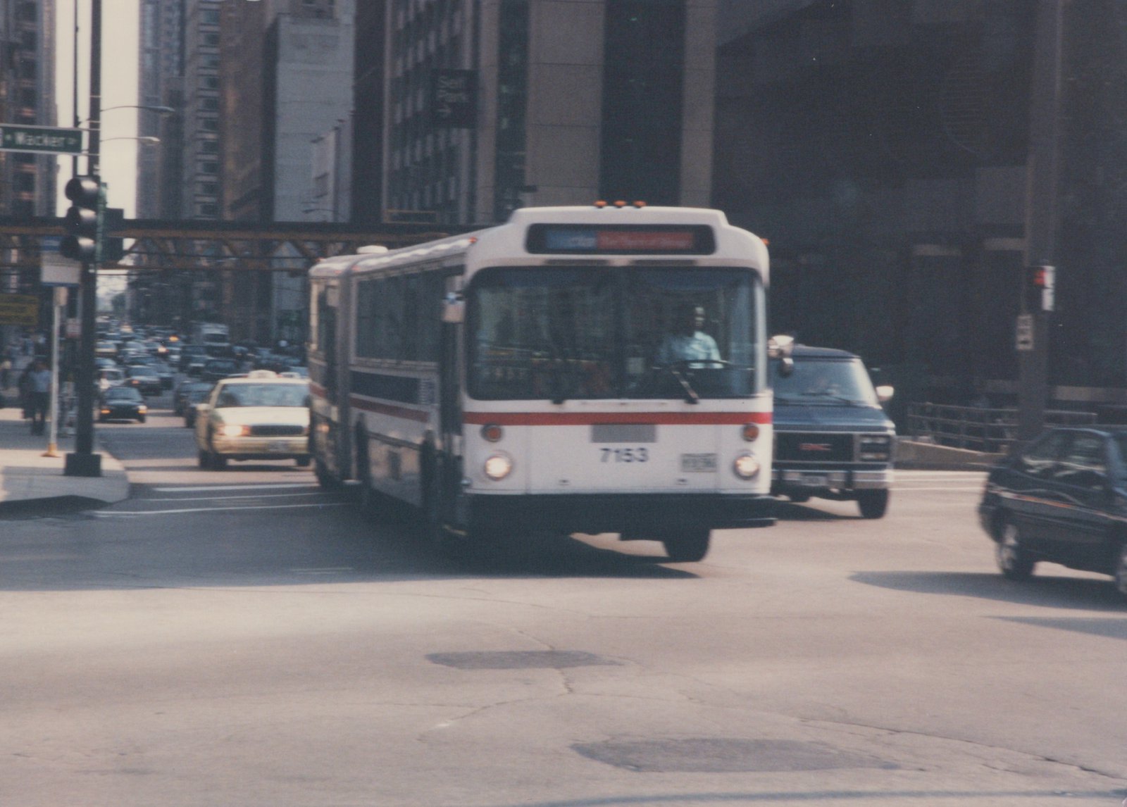 A white and red CTA bus, number 7153, is driving on a city street with other cars and an elevated structure in the background.