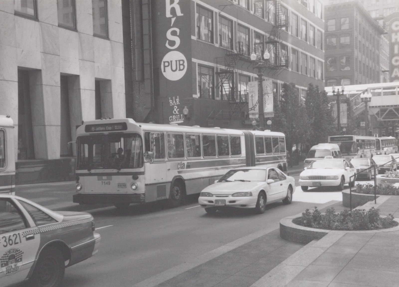A black and white photo of a CTA bus driving down a city street with cars and taxis, passing by buildings with signs for K's Pub and The Bar & Grill.