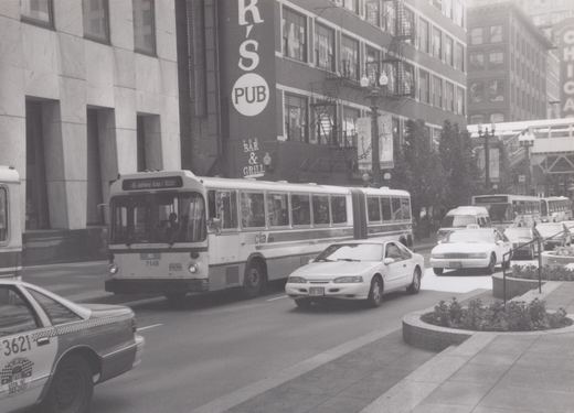 A black and white photo of a CTA bus driving down a city street with cars and taxis, passing by buildings with signs for K's Pub and The Bar & Grill.