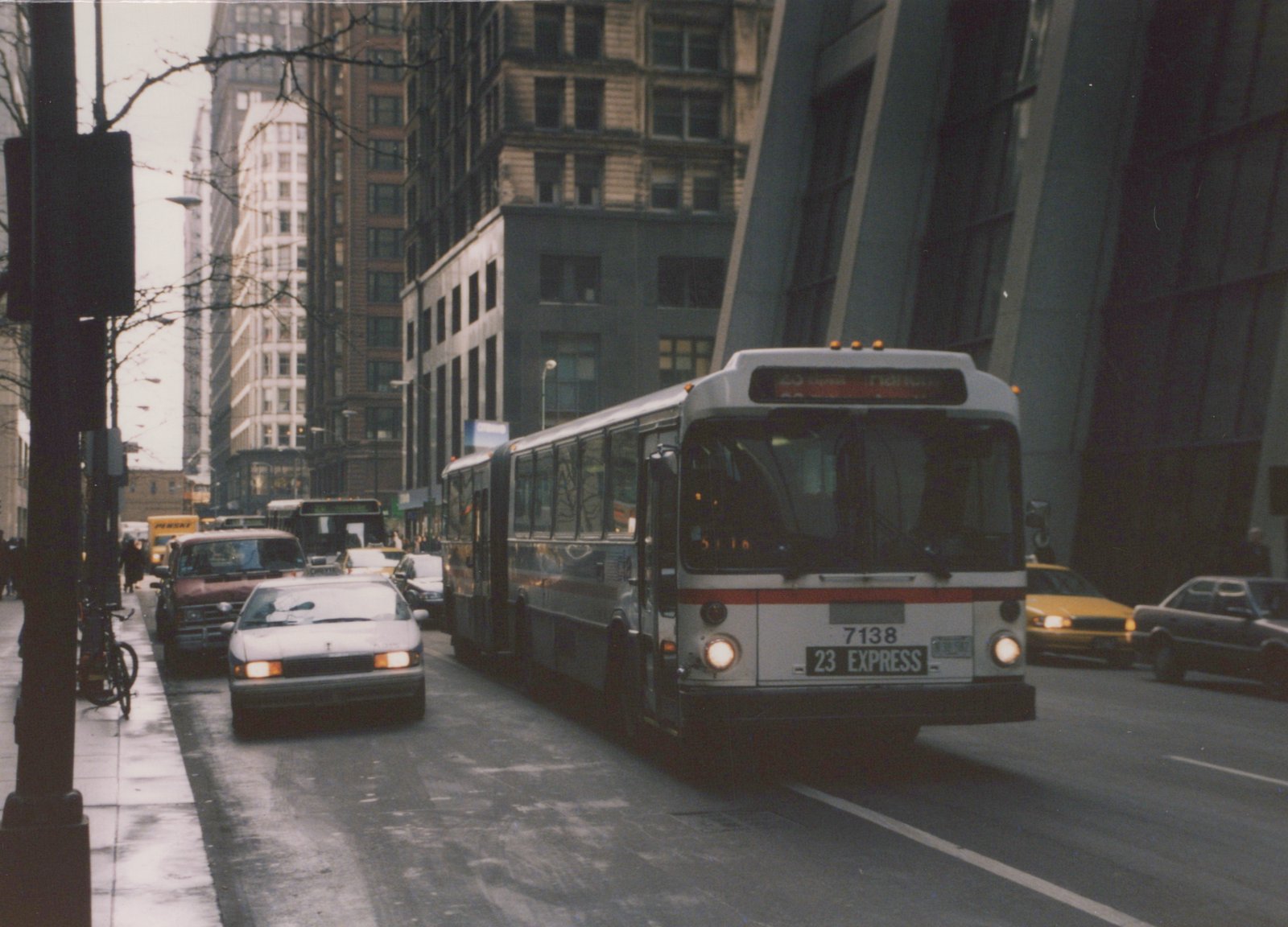 A white and red CTA bus marked 7138 on the 23 Express route is driving down a wet city street lined with tall buildings, with other cars and a taxi nearby.
