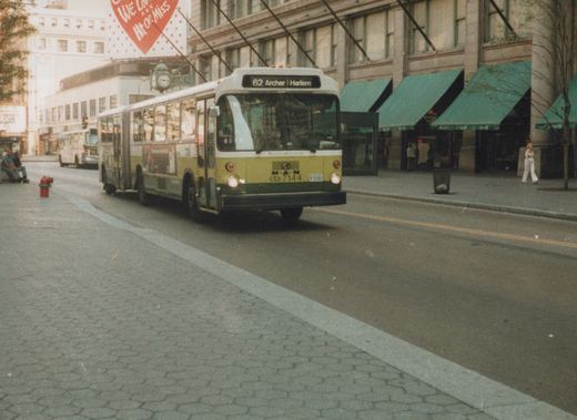 A green and white trolleybus with the number 7144 on the front drives down a city street lined with buildings and pedestrians.