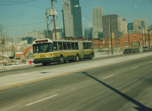 A green, yellow, and cream articulated bus with the number 7145 on the front is driving on a city street in winter, with tall buildings visible in the distance.