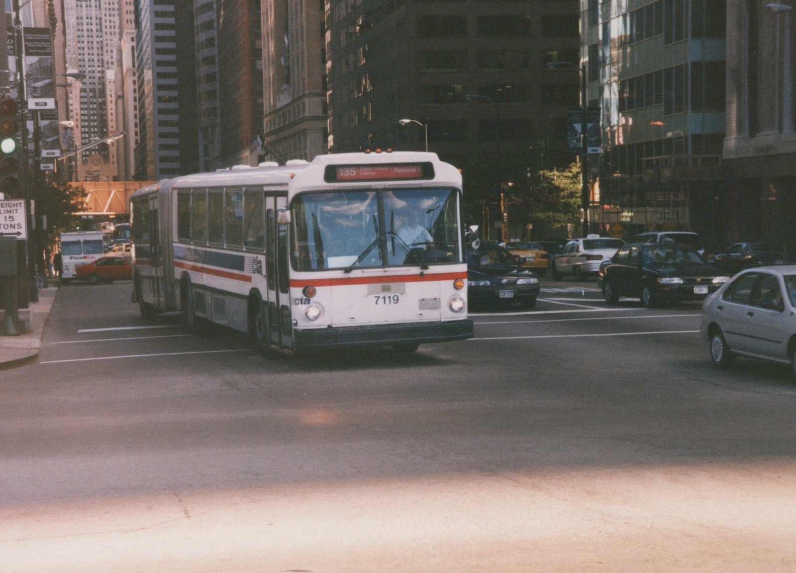 A white Chicago Transit Authority bus with red and blue stripes, number 7119, is driving on a busy city street with tall buildings in the background.