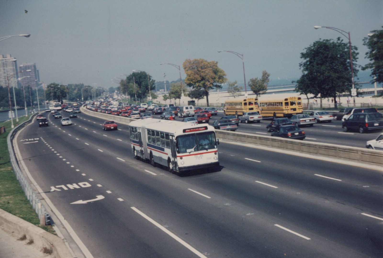 A white and blue articulated city bus drives on a multi-lane highway with many other cars, including two yellow school buses, on a hazy day with trees and buildings in the distance.