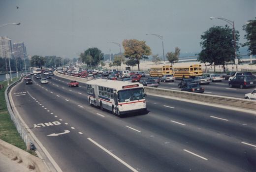 A white and blue articulated city bus drives on a multi-lane highway with many other cars, including two yellow school buses, on a hazy day with trees and buildings in the distance.