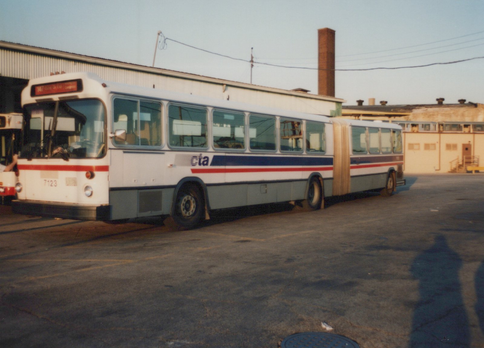 A white, blue, and red articulated bus with the number 7123 and CTA lettering is parked in a lot next to a large building, with a long shadow cast on the pavement.
