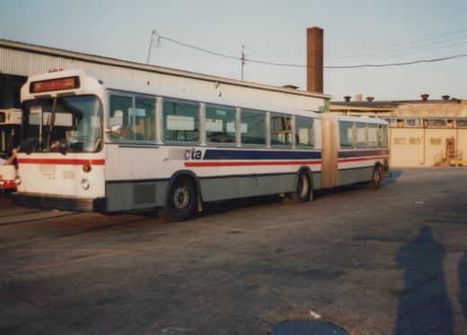 A white, blue, and red articulated bus with the number 7123 and CTA lettering is parked in a lot next to a large building, with a long shadow cast on the pavement.