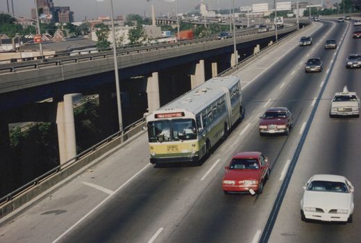 An elevated, angled view of a green and white articulated bus driving on a highway with several cars, with an overpass and city buildings in the background.