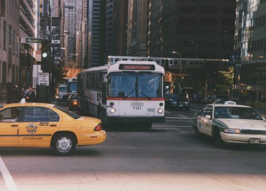 A white and red CTA bus drives down a street in Chicago, with a yellow taxi cab and an elevated train visible in the background.