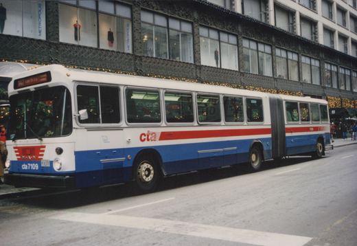A white, red, and blue articulated bus from the Chicago Transit Authority, numbered 7109, is parked on a city street during the winter holidays, with storefronts and pedestrians visible in the background.