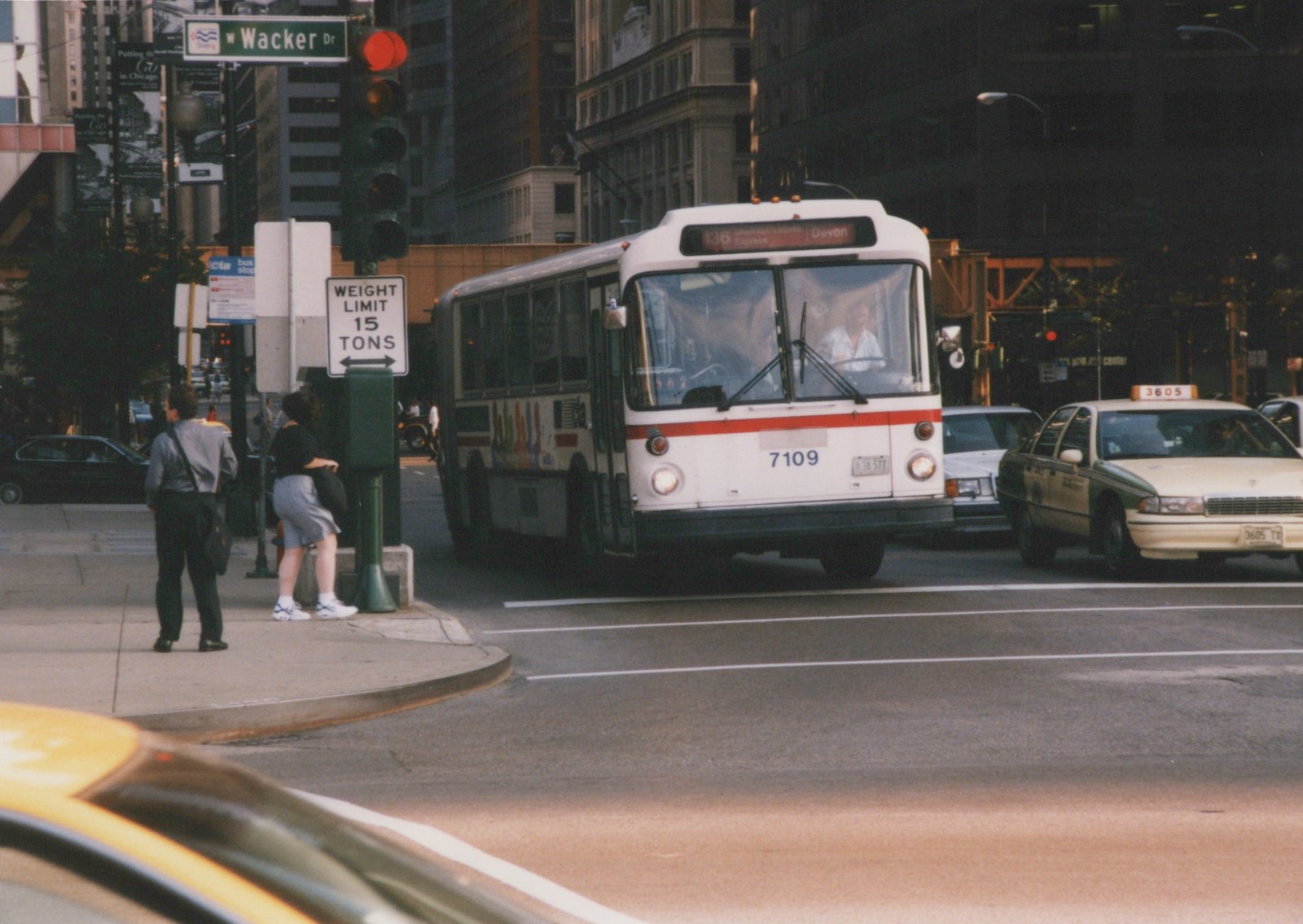A white and red CTA bus with the number 7109 on its front, stopped at an intersection on Wacker Drive in Chicago, with pedestrians and taxis nearby.