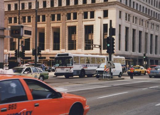 A white CTA bus with red and blue stripes, marked 'Not in Service', is stopped at a city street intersection with traffic lights, surrounded by taxis and other cars; a traffic officer directs traffic nearby.