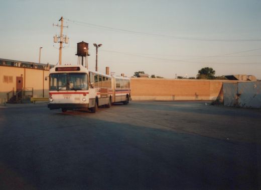 A white CTA articulated bus with red and blue stripes, numbered 7107, is parked in a lot with a brick wall and a water tower in the background.