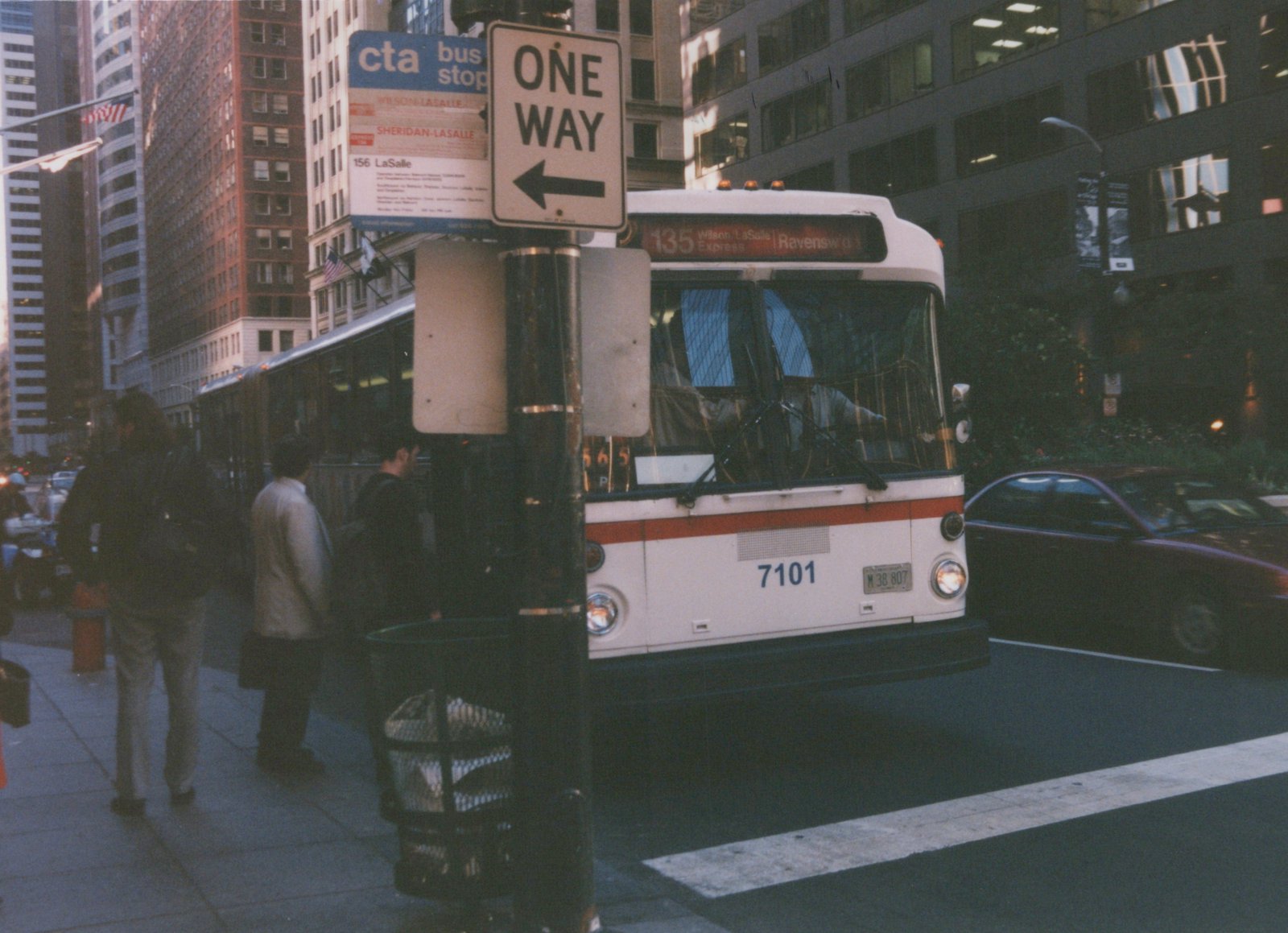 A white and orange Chicago Transit Authority bus, number 7101, is stopped at a bus stop on a city street with tall buildings in the background.