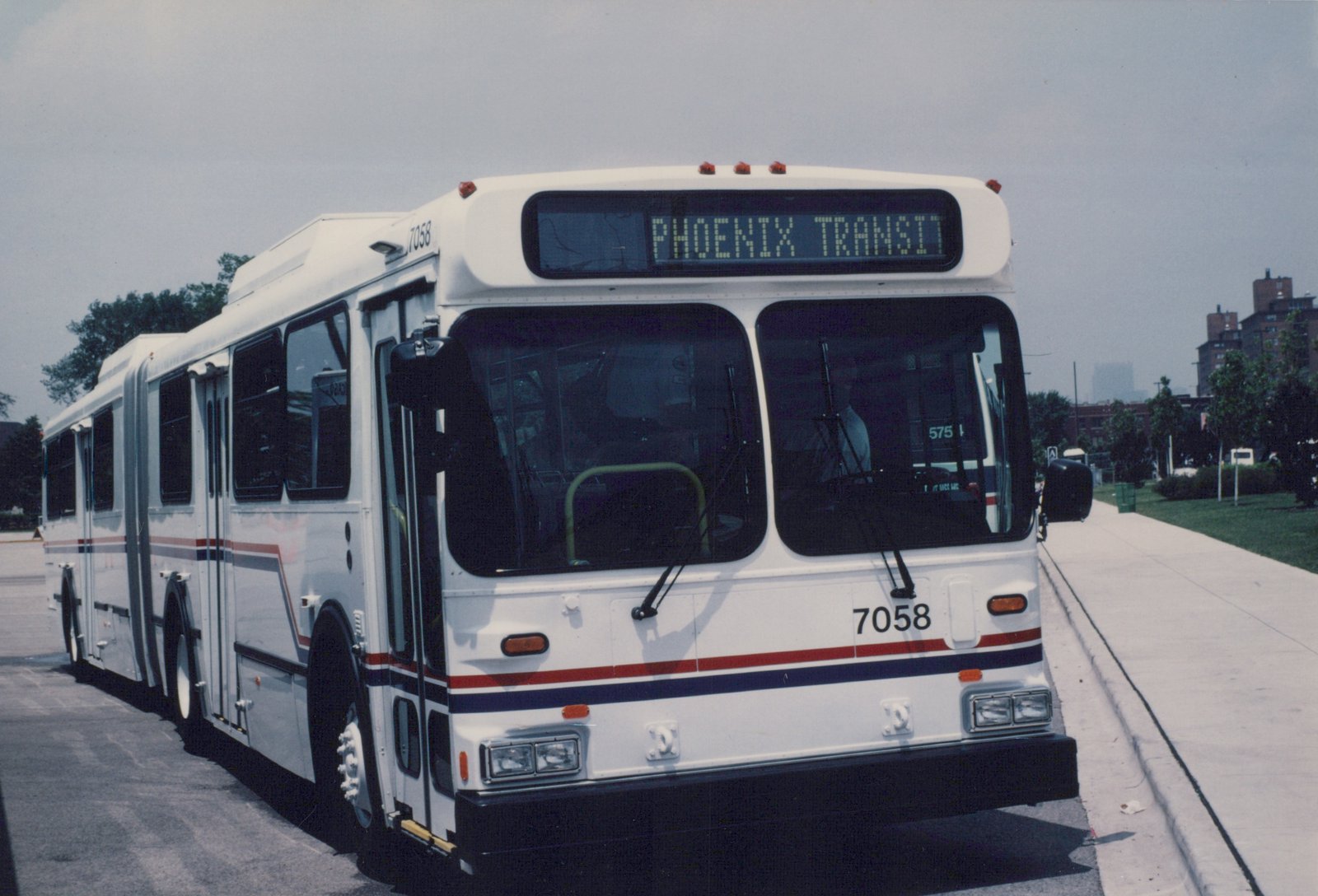 A white articulated bus with red and blue stripes, marked Phoenix Transit and number 7058, is parked on a city street with trees and buildings in the background.