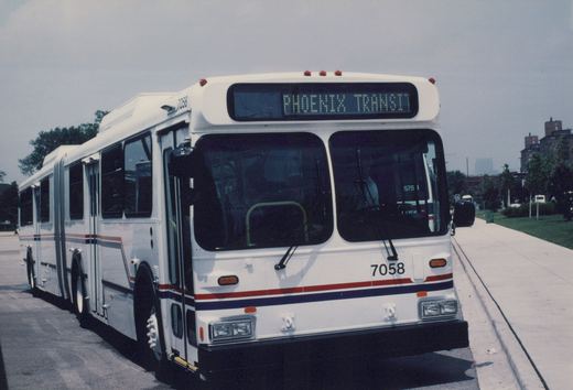 A white articulated bus with red and blue stripes, marked Phoenix Transit and number 7058, is parked on a city street with trees and buildings in the background.
