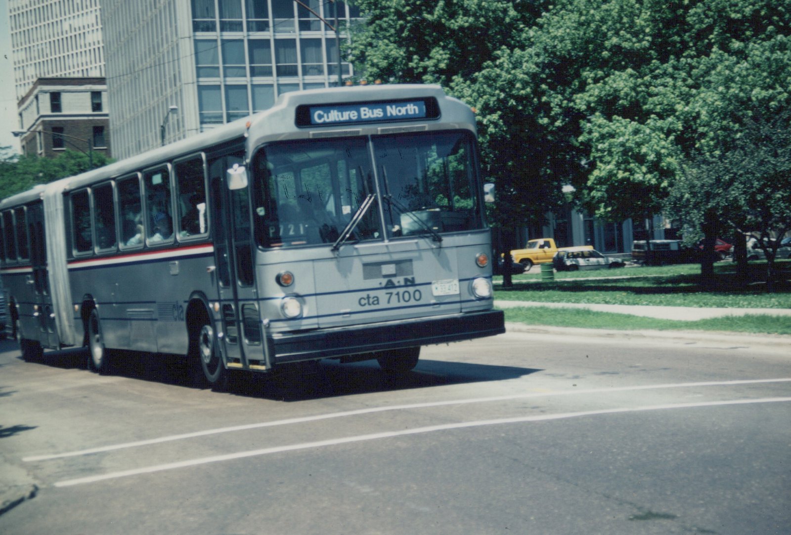 A silver articulated city bus with blue and red stripes drives on a multi-lane road; trees and buildings are in the background.