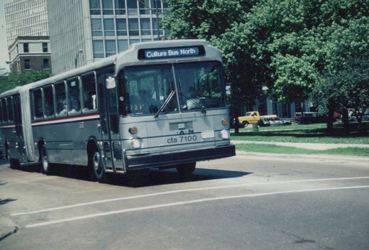 A silver articulated city bus with blue and red stripes drives on a multi-lane road; trees and buildings are in the background.