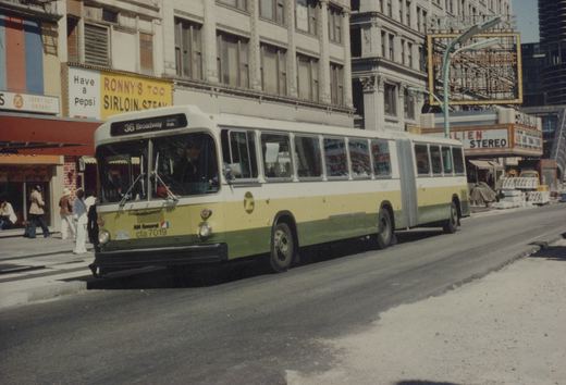 A white, yellow and green articulated CTA bus on a city street with people on the sidewalk and movie theater marquees in the background.