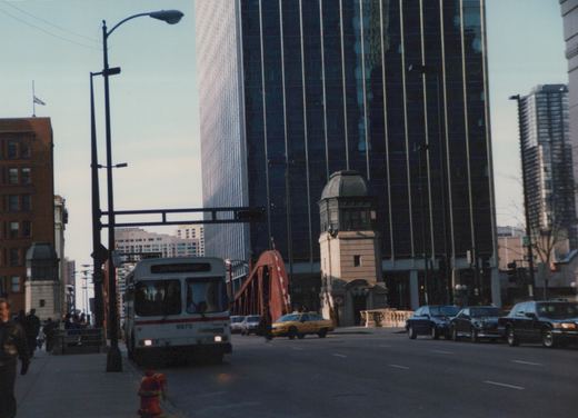 Front view of CTA bus number 9970, a white and red motor bus, on a downtown Chicago street near a drawbridge with pedestrians and cars nearby.