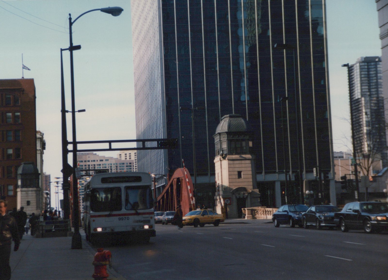 Front view of CTA bus number 9970, a white and red motor bus, on a downtown Chicago street near a drawbridge with pedestrians and cars nearby.