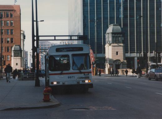 A white Chicago Transit Authority bus with the number 9971 and route 24 Wentworth displayed on its front, driving on a city street with pedestrians and tall buildings in the background.