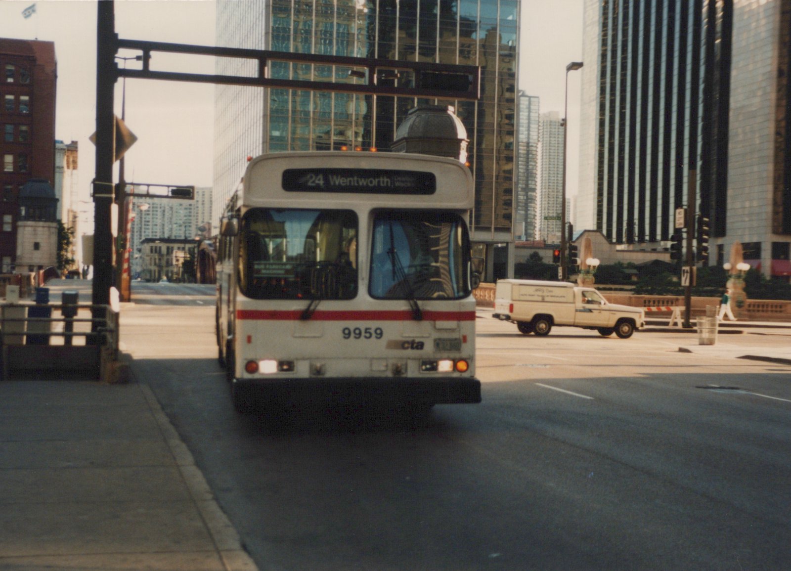 Front view of a white CTA bus with a red stripe, number 9959, on route 24 Wentworth, driving down a city street with tall buildings in the background and a pickup truck nearby.