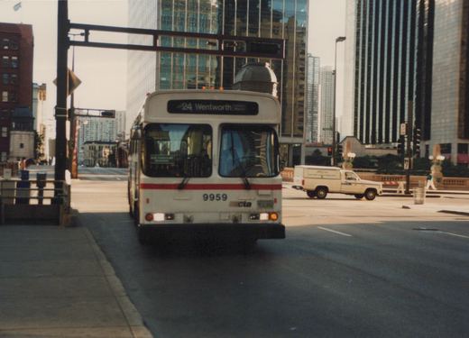 Front view of a white CTA bus with a red stripe, number 9959, on route 24 Wentworth, driving down a city street with tall buildings in the background and a pickup truck nearby.