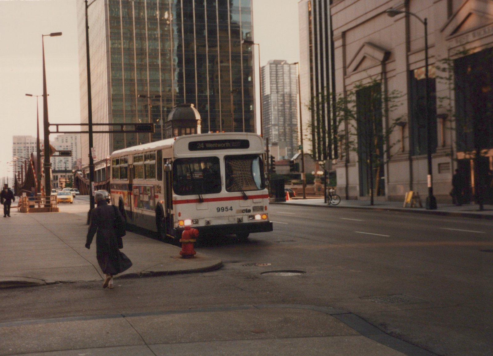 A white and red Chicago Transit Authority bus with the number 9954 and route 24 Wentworth on its sign is stopped on a city street, with a pedestrian walking on the sidewalk and tall buildings behind it.