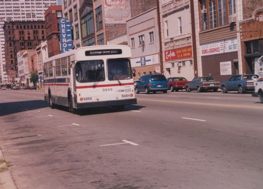 A white and red CTA bus with the number 9949 on its front, driving down a city street with parked cars and brick buildings in the background.