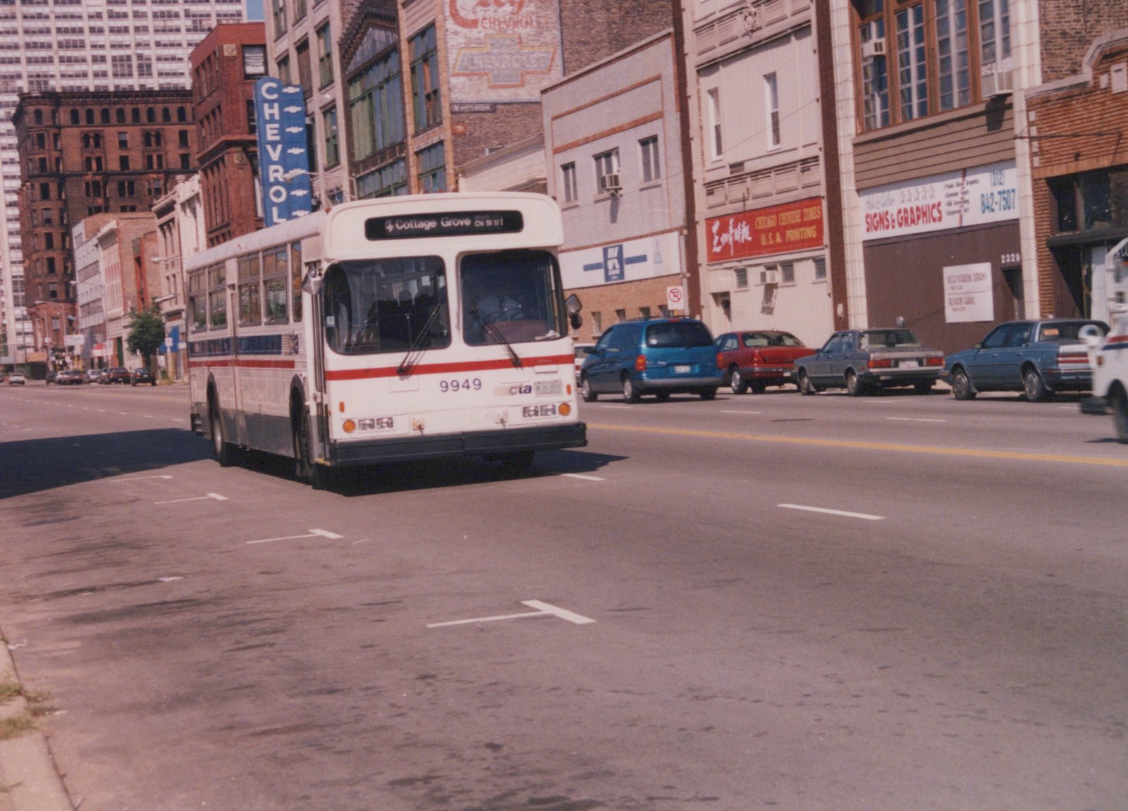 A white and red CTA bus with the number 9949 on its front, driving down a city street with parked cars and brick buildings in the background.