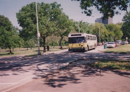 A yellow, white, and green CTA bus, number 9944, drives down a shaded street lined with trees and parked cars on a clear day.