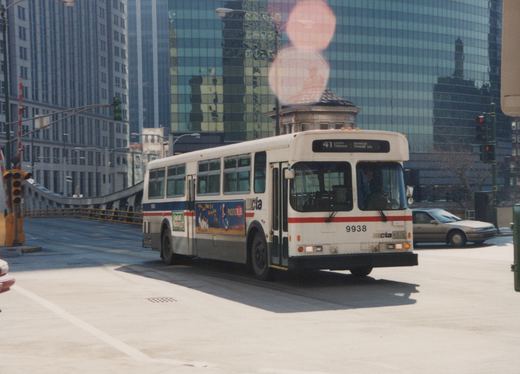 A white, red, and blue CTA bus with the number 9938 and route 41 sign is driving on a city street near a bridge with tall buildings in the background.