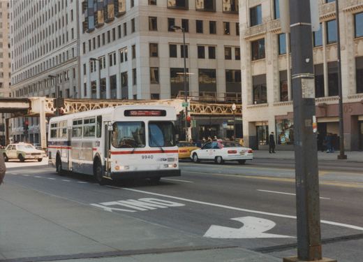 A white CTA bus with the number 9940 is driving on a city street with tall buildings and taxis in the background, at the corner of Wells and Wacker.