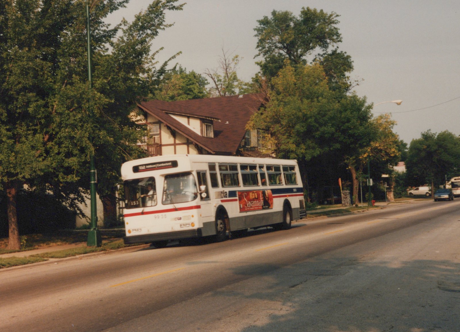 A white CTA bus with red and blue stripes, number 9935, drives down a tree-lined street with a Tudor-style house in the background.