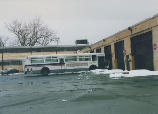 A white and blue Chicago Transit Authority bus, number 9930, is parked in a snowy lot next to a brick garage, with two people near its door.