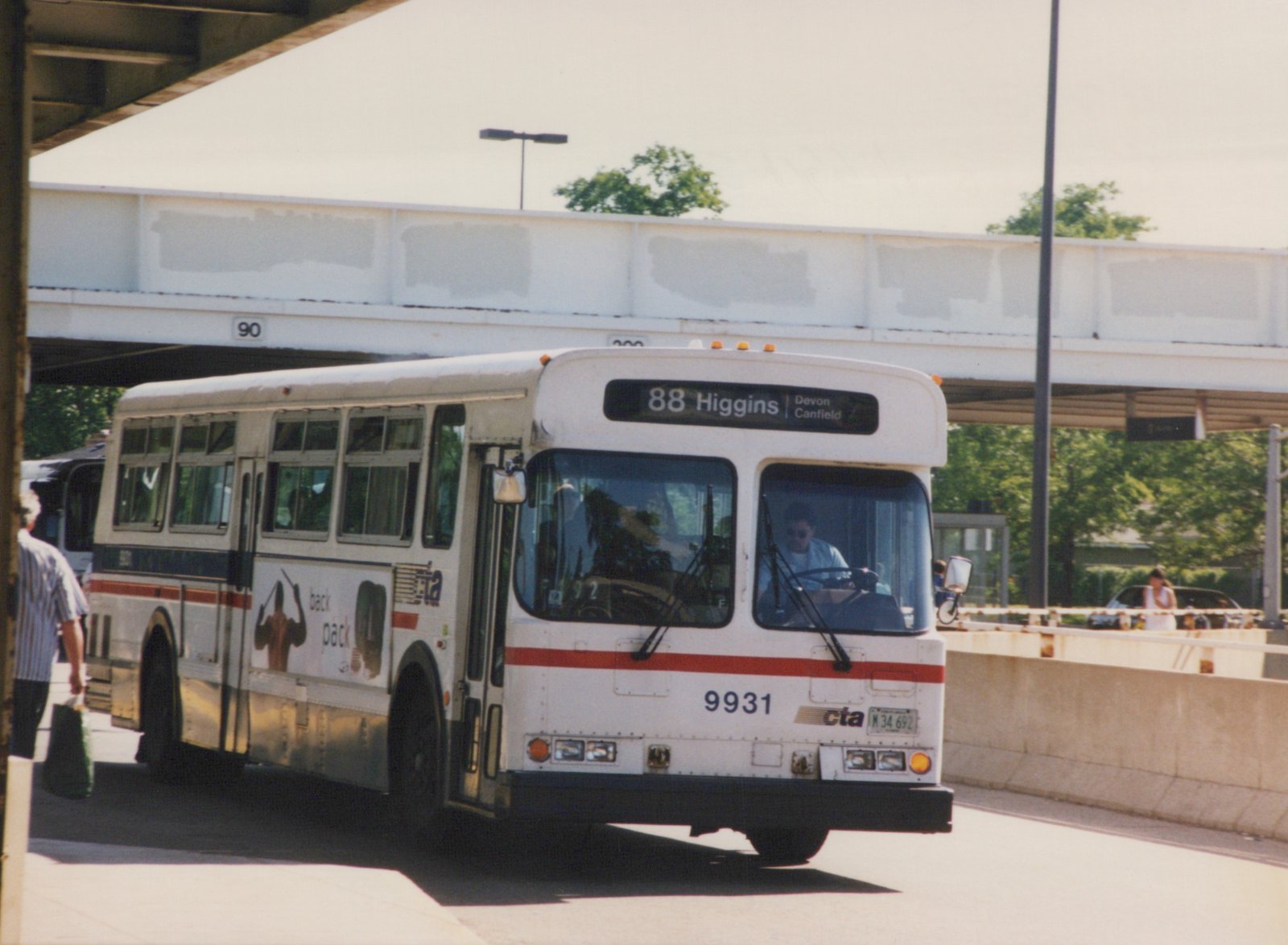 A white CTA bus with red and blue stripes, number 9931, is driving on a road under an overpass, with a driver visible and pedestrians nearby.