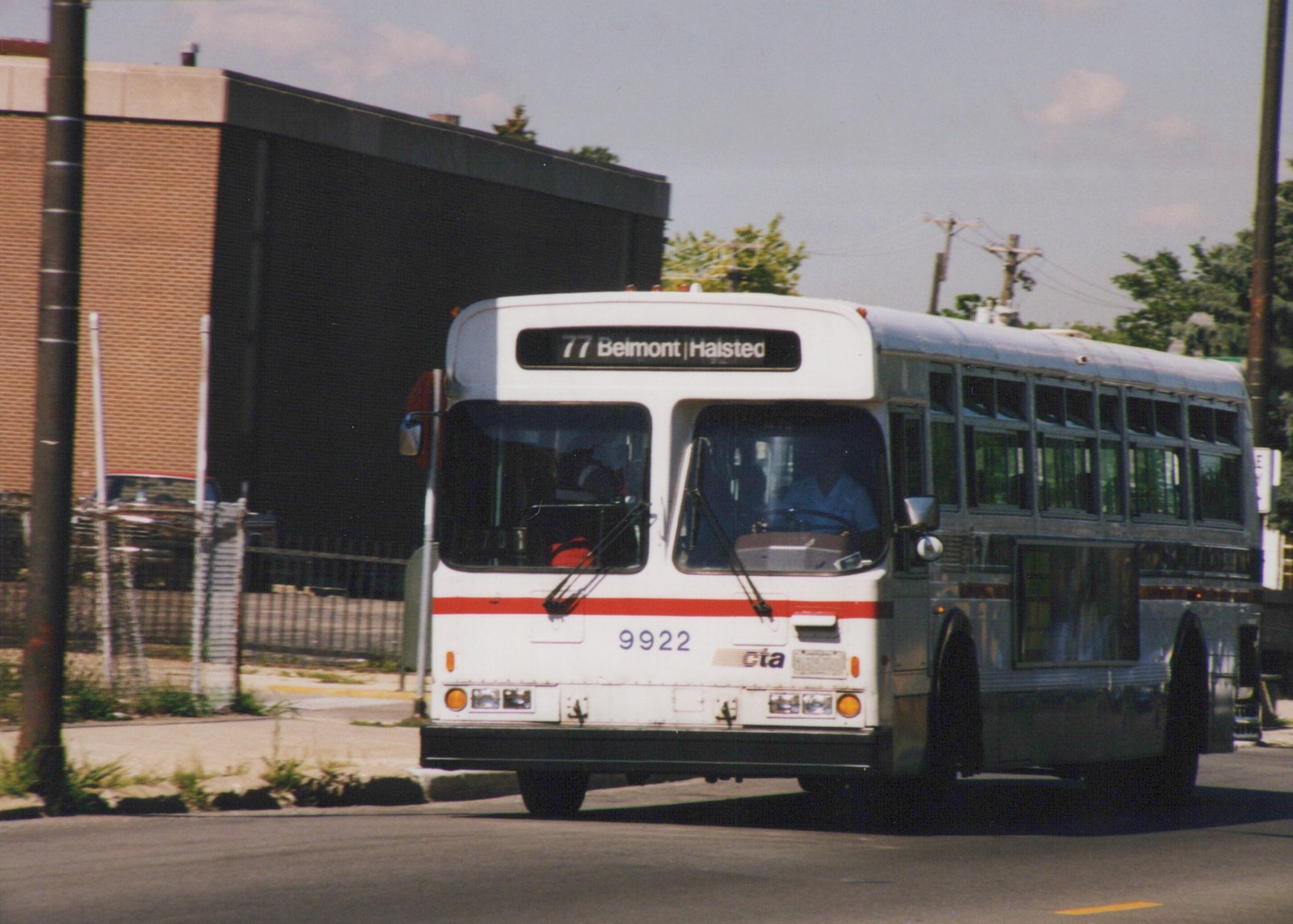 A white and red Chicago Transit Authority bus, number 9922, on route 77 Belmont/Halsted, driving on a street with a brick building in the background on a sunny day.
