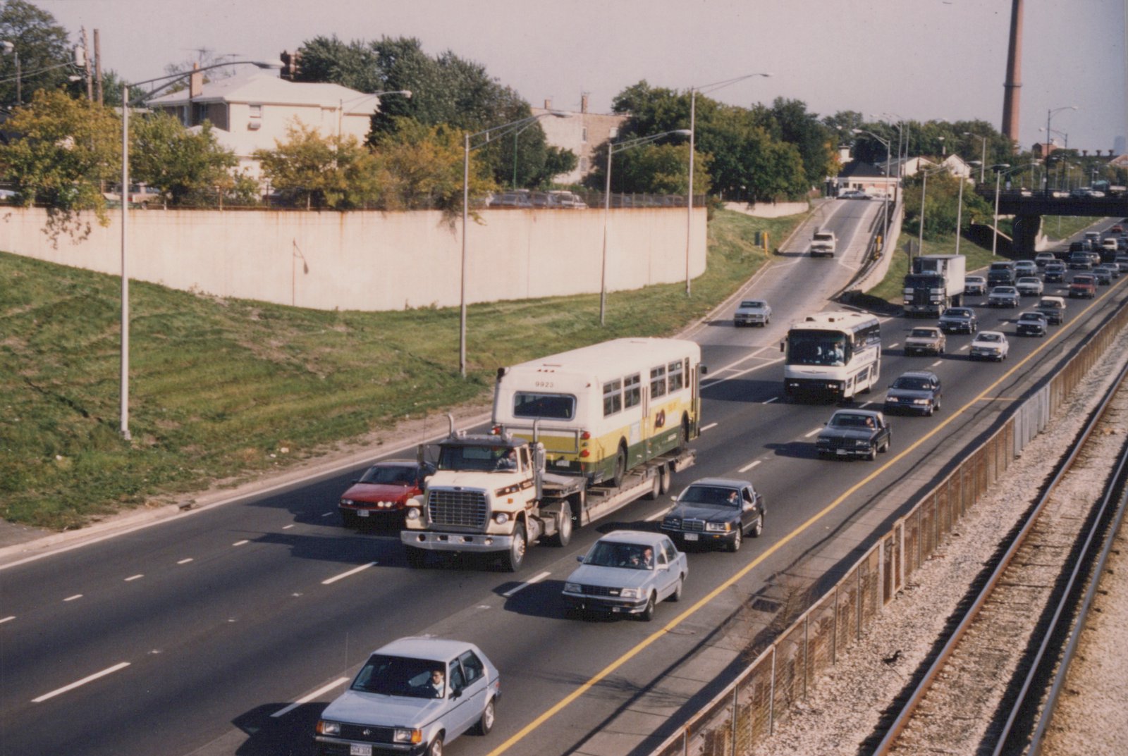 A white and green Chicago Transit Authority bus is on a flatbed truck on a multi-lane highway, surrounded by 1980s cars and a motor coach, with railroad tracks visible on the right.