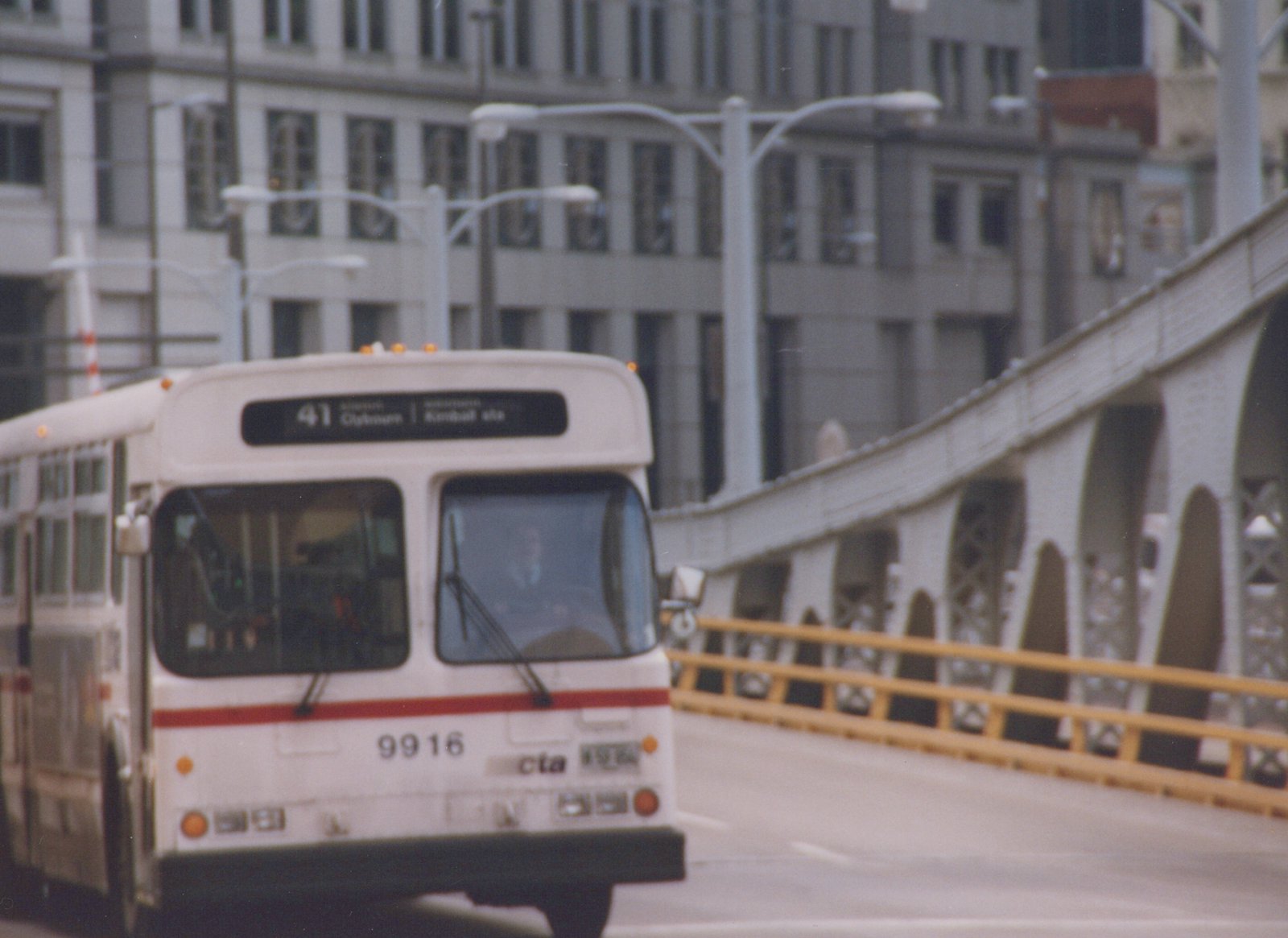 A white Chicago Transit Authority bus with red stripe, number 9916, is shown from a front three-quarter view crossing a bridge, with a city building in the background.