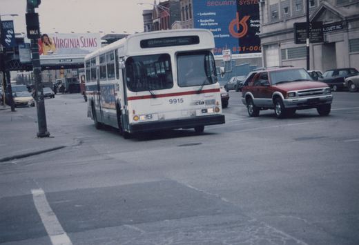 A white CTA bus with the number 9915 on the front is driving down a city street, with a red SUV and other cars nearby on an overcast day.