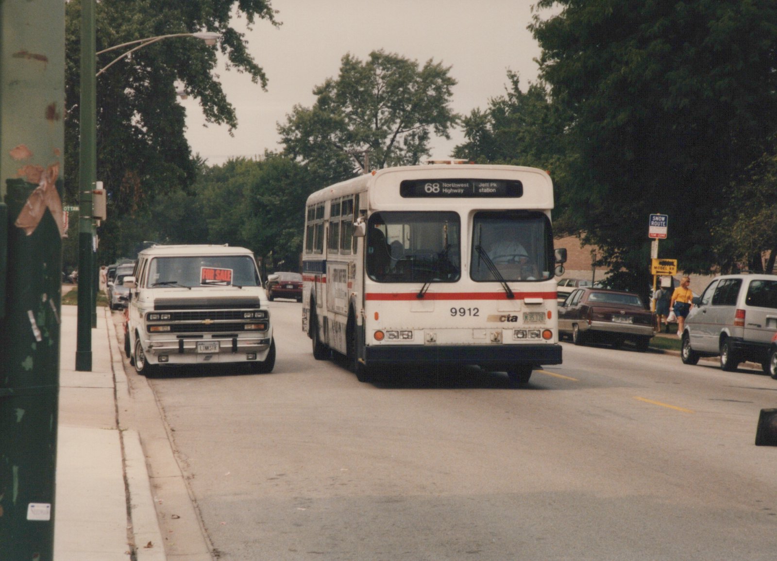 A white Chicago Transit Authority bus with red and gold stripes, numbered 9912, drives down a street lined with parked cars and trees, with a destination sign reading '68 Northwest Highway Jeff Pk station'.