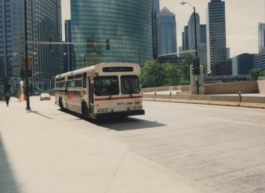 A white Chicago Transit Authority bus with red and blue stripes, numbered 9910, is driving on a paved road with tall city buildings in the background. A yellow taxi cab is visible behind the bus, and a person is walking on the sidewalk.