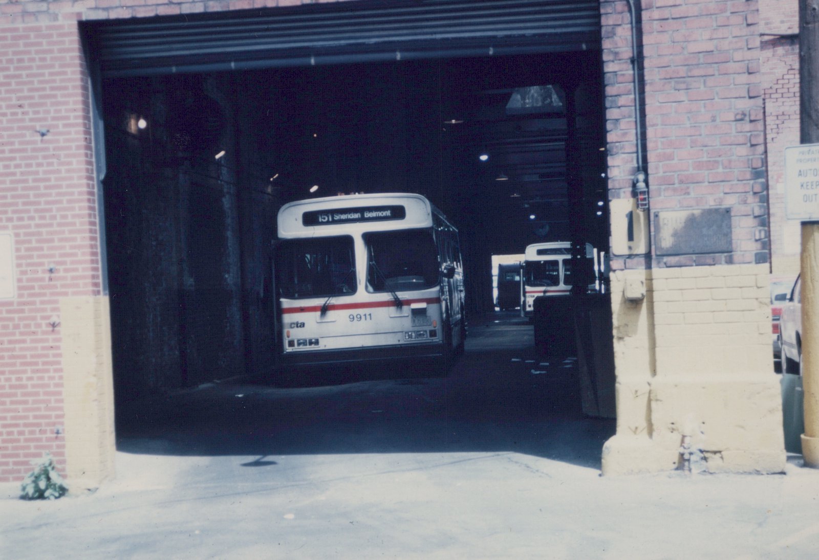Front view of a white and red CTA bus, fleet number 9911, parked in a brick garage with snow on the ground outside.