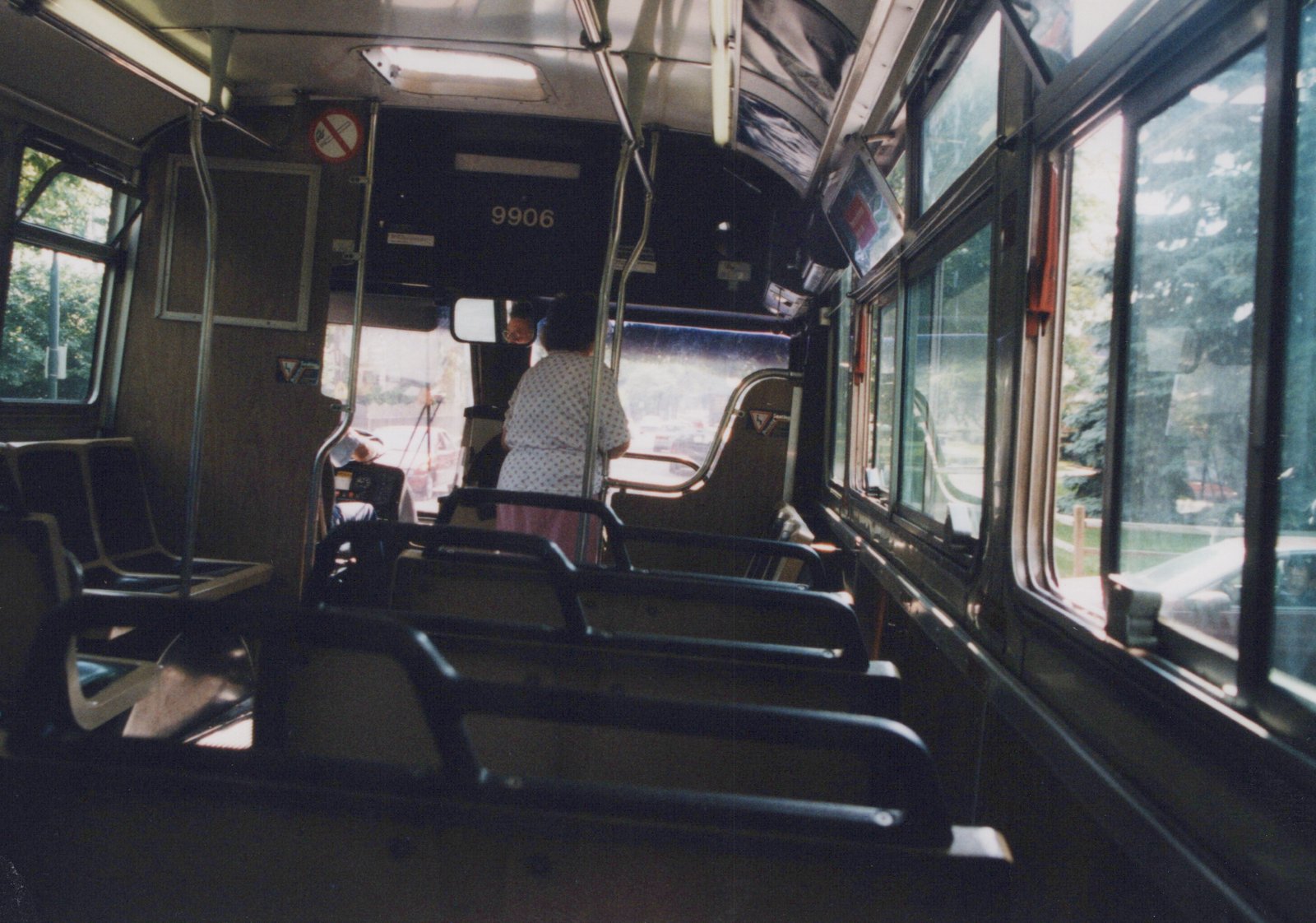 The interior of a city bus, showing rows of seats, a passenger standing, and the driver, with the number 9906 visible on the front bulkhead and trees visible through the windows.