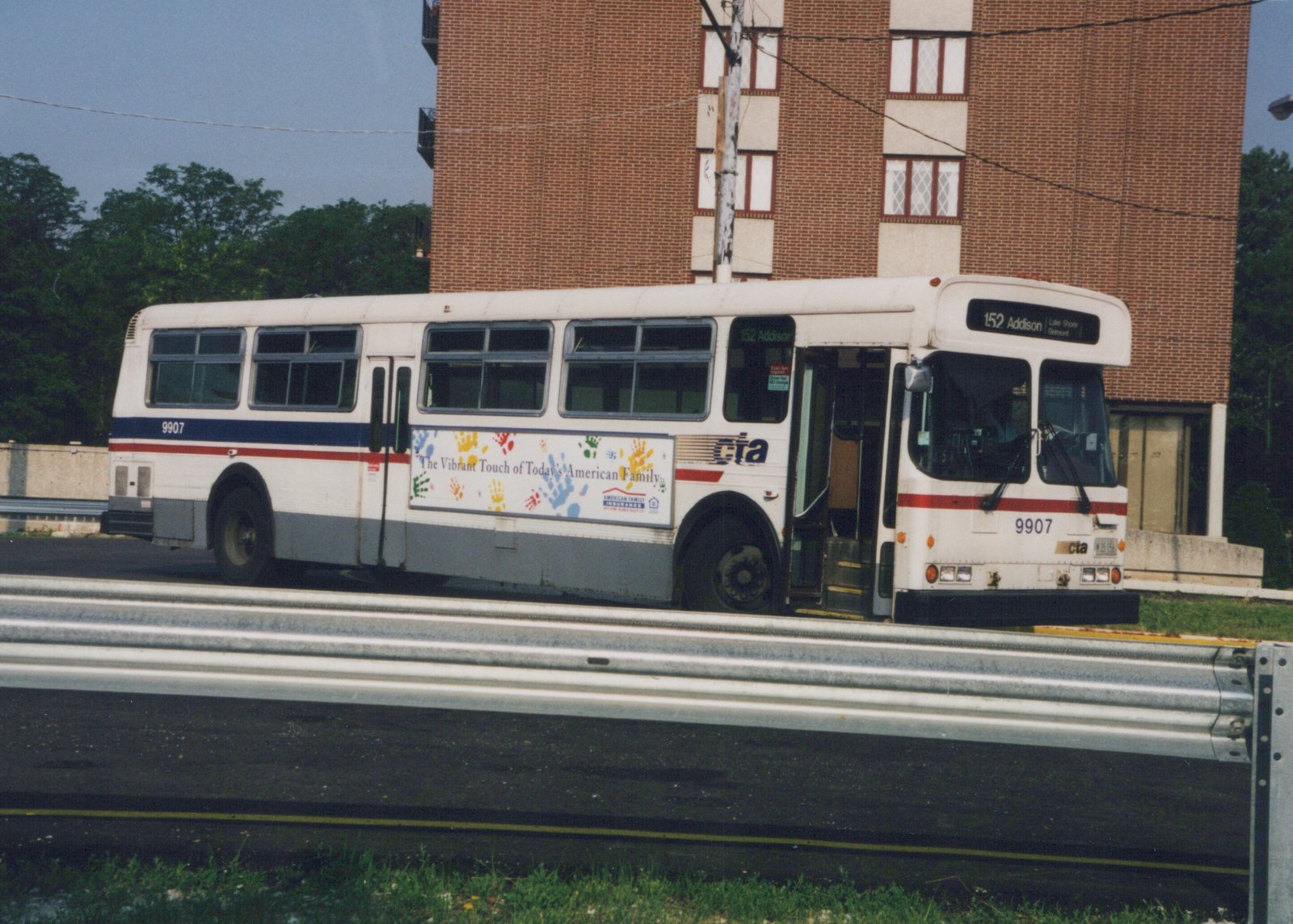 A white CTA motor bus with blue and red stripes, numbered 9907, is parked on an expressway median. The destination sign reads '152 Addison.' The side features an advertisement for American Family Insurance.